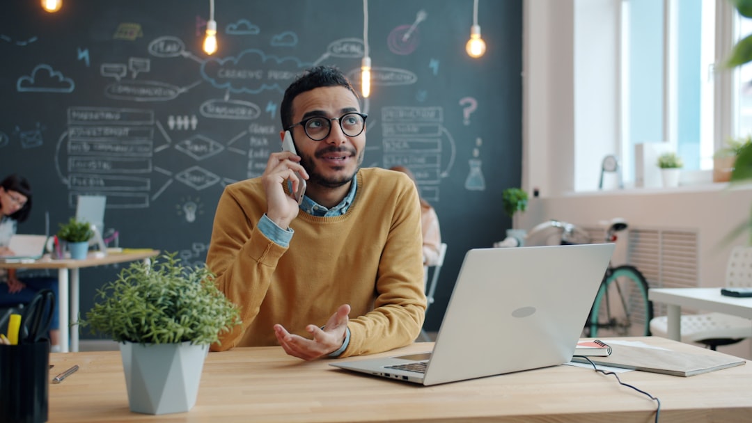 Man talking on his phone at a desk with a laptop open, representing everyday use of an AI assistant powered by an LLM