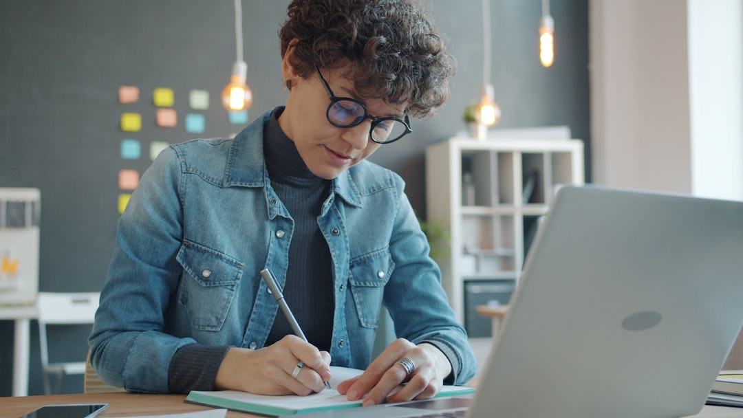 Woman with glasses writing in a notebook at a desk, representing studying for a certification